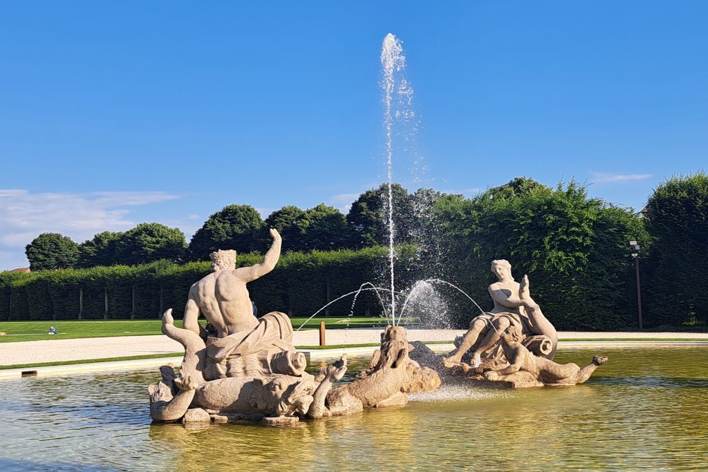 Artistic fountain featuring statues in a pond with water jets, set against green trees and a blue sky.